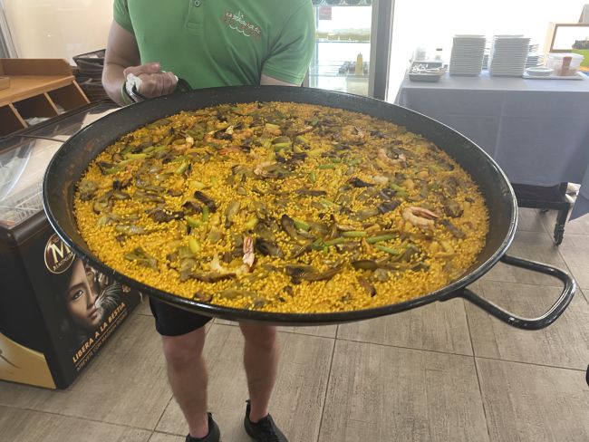 Ein Mann hält eine große Pfanne mit Paella hoch. Viel Reis, eine gelbliche Soße, Fleisch und Fischstücke darin.