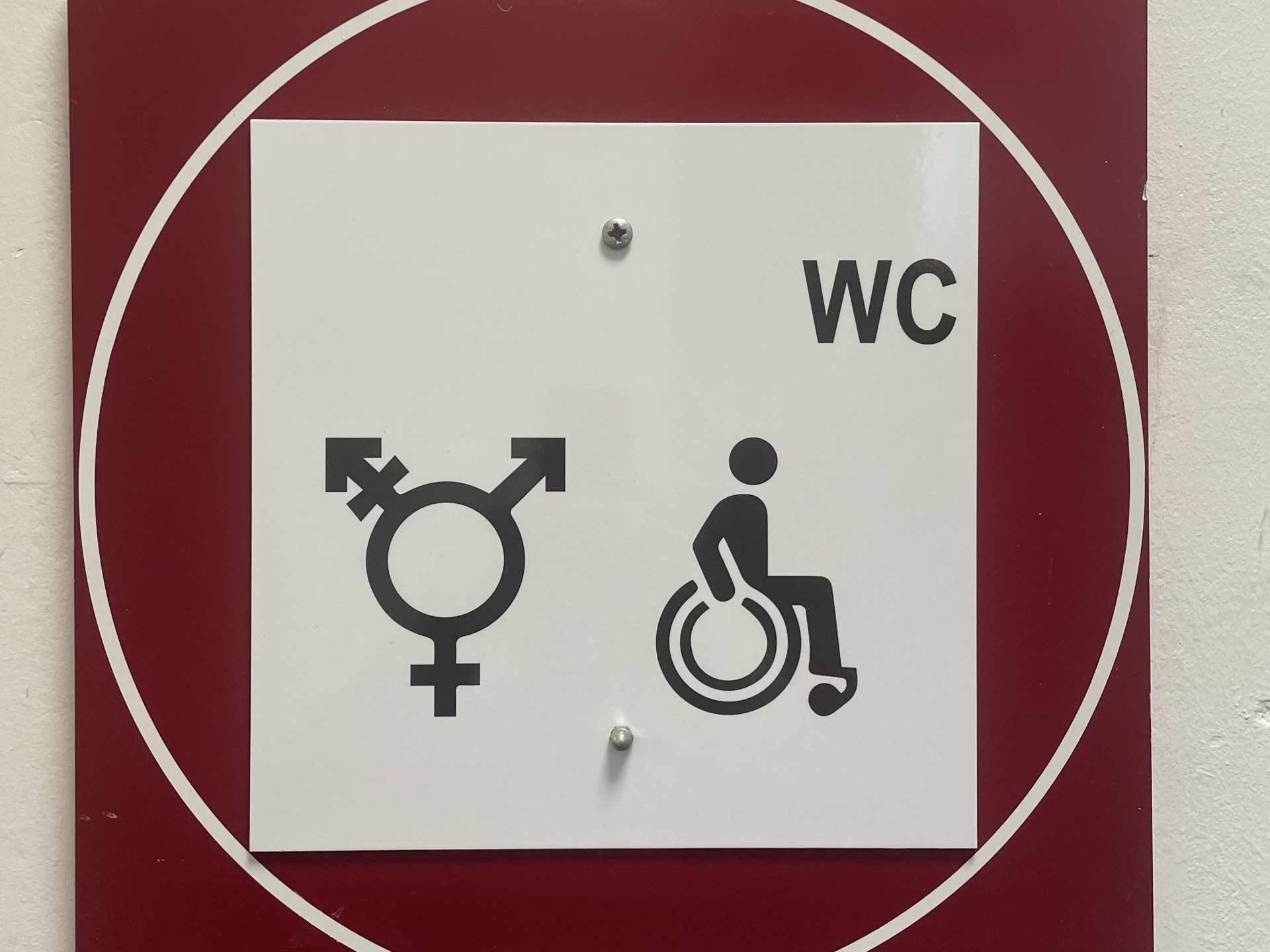Ein weißes quadratisches Schild in einem weißen Kreis auf einem roten Brett. Auf dem Schild ist rechts eine Person im Rollstuhl abgebildet. Links ein Kreis mit einem weiblich Zeichen nach unten, einem männlich Zeichen nach schräg rechts oben und einem Pfeil mit durchgestrichenem Strich.