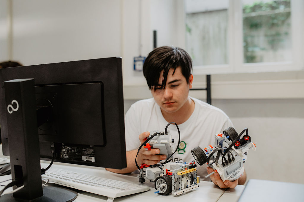 Ein Schüler sitzt vor einem Monitor und hält zwei Bauteile für einen Roboter in der Hand.