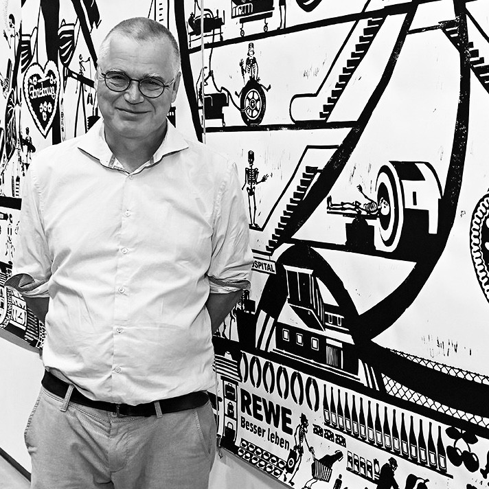 Flrorien Haas, Ein Künstler mit kurzen Haaren und Brille vor einer Wand mit schwarzen Zeichnungen.