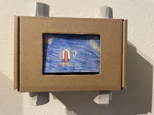 Eine Pappbox mit "Fenster" an beiden Seiten gibt es Stäbe zum drehen, damit so etwas wie ein Kino entsteht. Auf dem Bild im Fenster ist eine organge Rakete auf blauem Hnitergrund