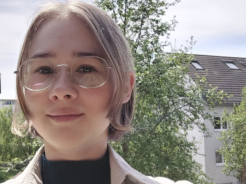 Eine junge Frau mit blonden, nach hinten zussammengebundenen Haaren, einem schwarzen Pullover und einer beigen Jacke. Im Hintergrund sind Häuserreihen und Bäume.
