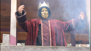Eine goldene Krone auf dem Kopf und eine goldene Gesichtsmaske tragend steht eine Person im purpurnen Gewand mit ausgestreckten Armen auf einem Balkon.