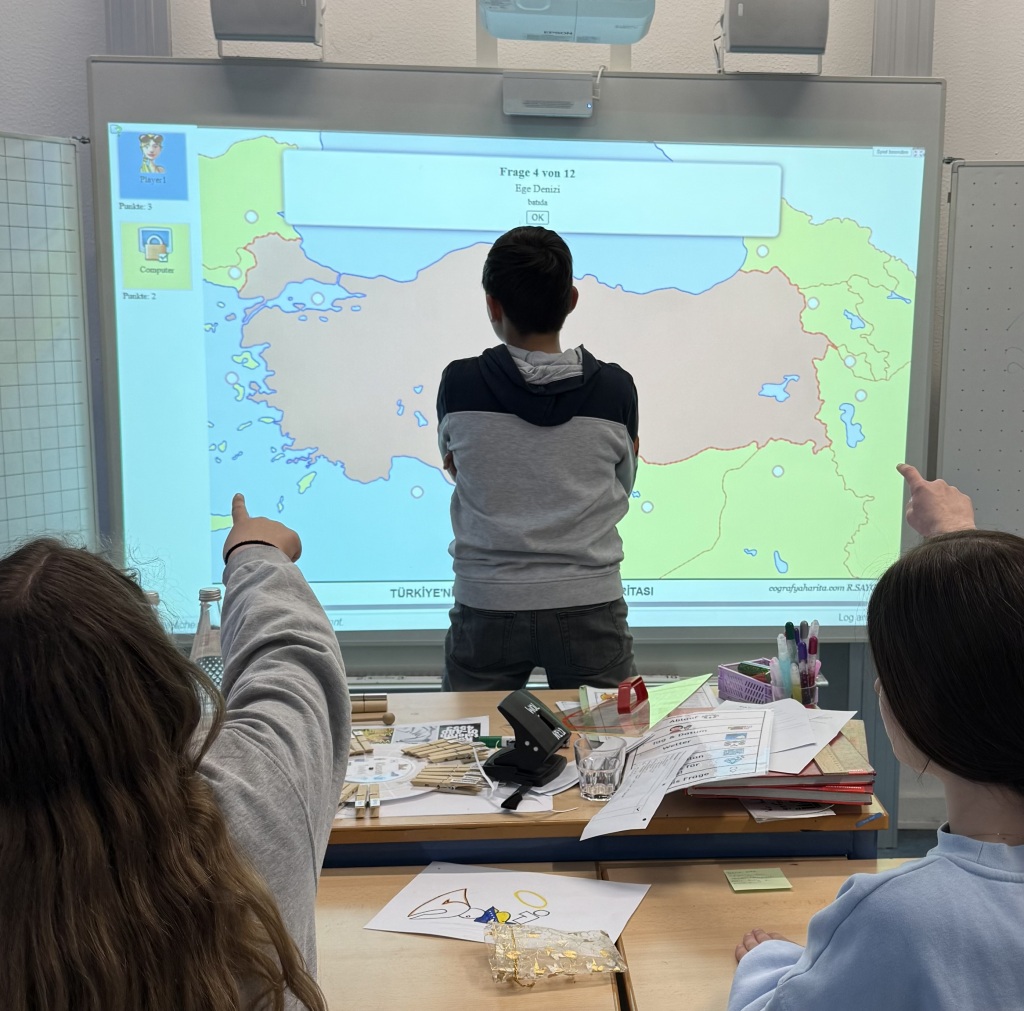 Ein Schüler steht vor einer Projektion auf der Wand mit der Karte der Türkei. Im Vordergrund sitzen 2 Schülerinnen.