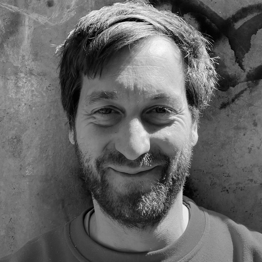 Ein Männer lächelt in die Camera, er hat einen Mittelscheitel und einen Vollvart und ein spitzes Kinn.