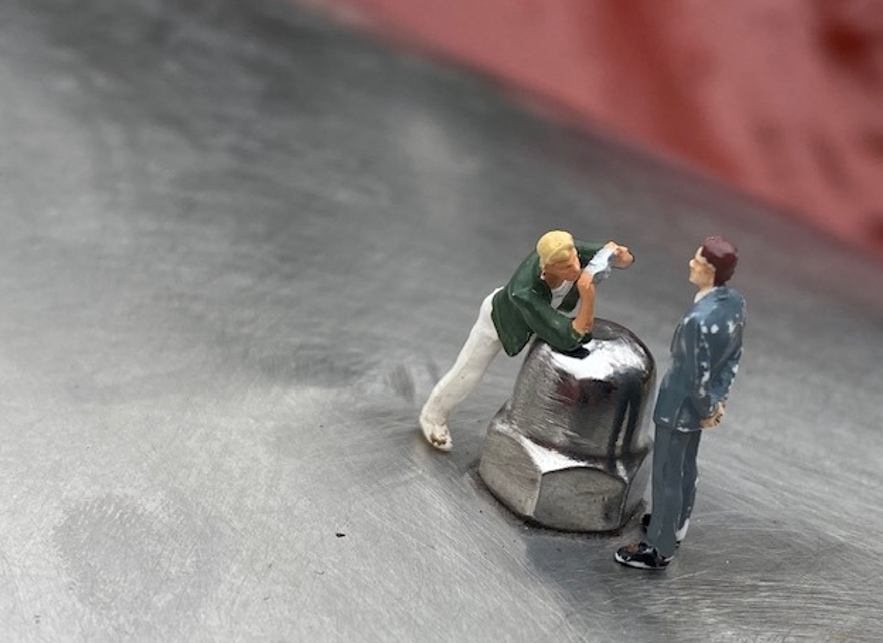Auf einer metallenen Fläche lehnt eine H0 Figur und redet mit einer anderen Figur im Anzug. Sie lehnt auf einerm großen Schraubenkopf.