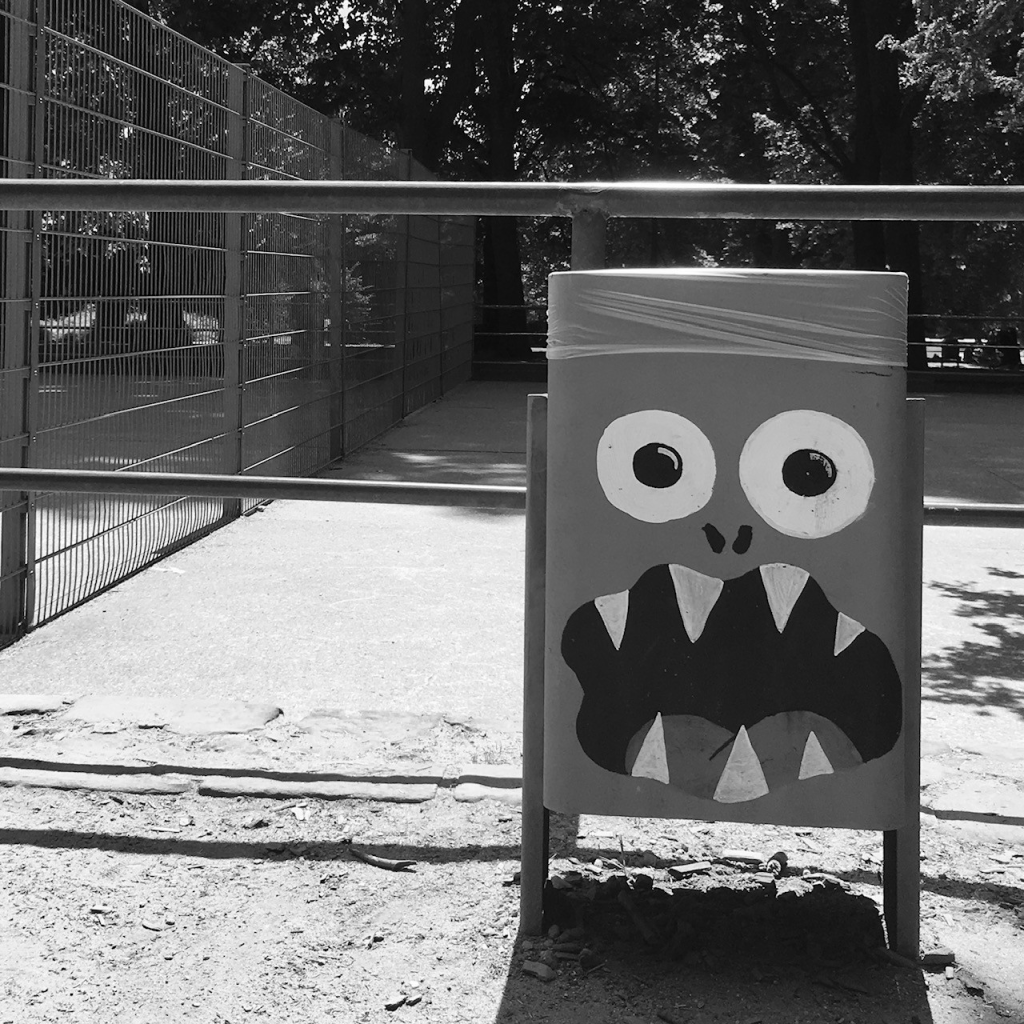 Ein Mülleiner auf einem Kinderspielplatz auf dem ein Gesicht mit weit offenen Augen und großem offenen Mund mit scharfen Zähnen gemalt ist.