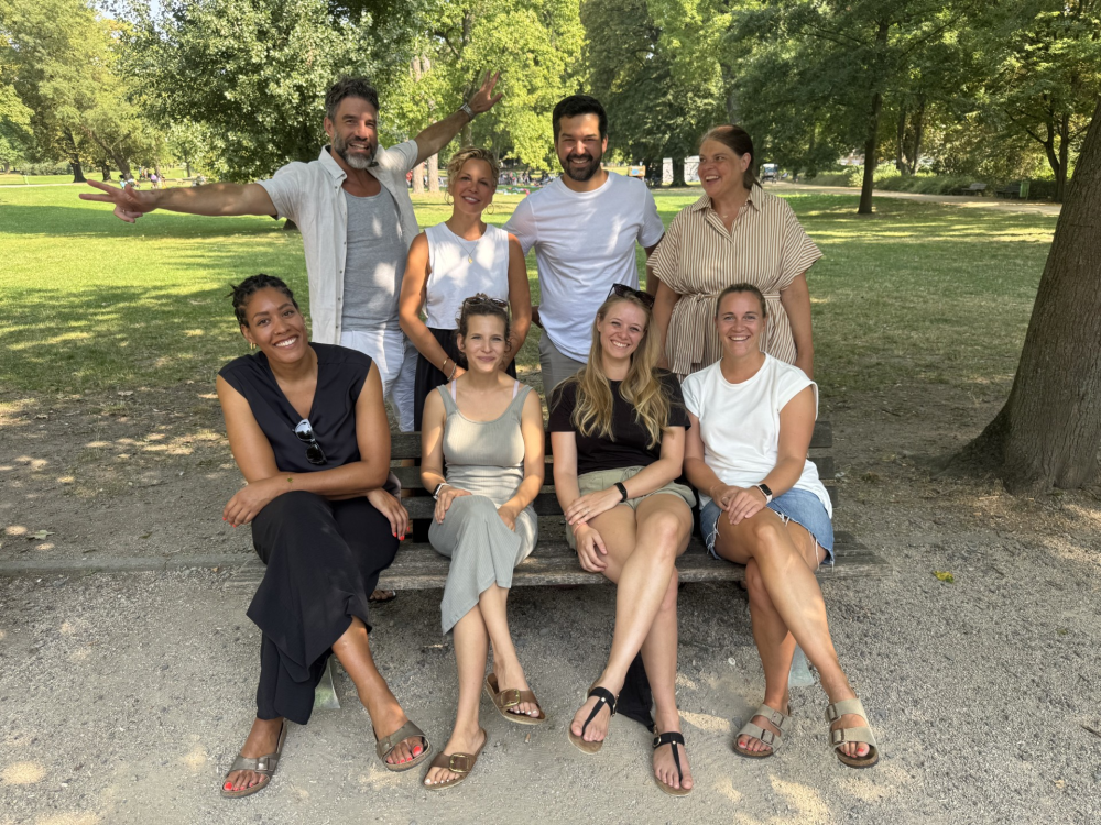 Eine Gruppe von Erwachsenen im Park. 4 Frauen sitzen auf der Bank. 2 Männer und 2 Frauen stehen hinter der Bank. sie tragen leichte Sommerkleidung