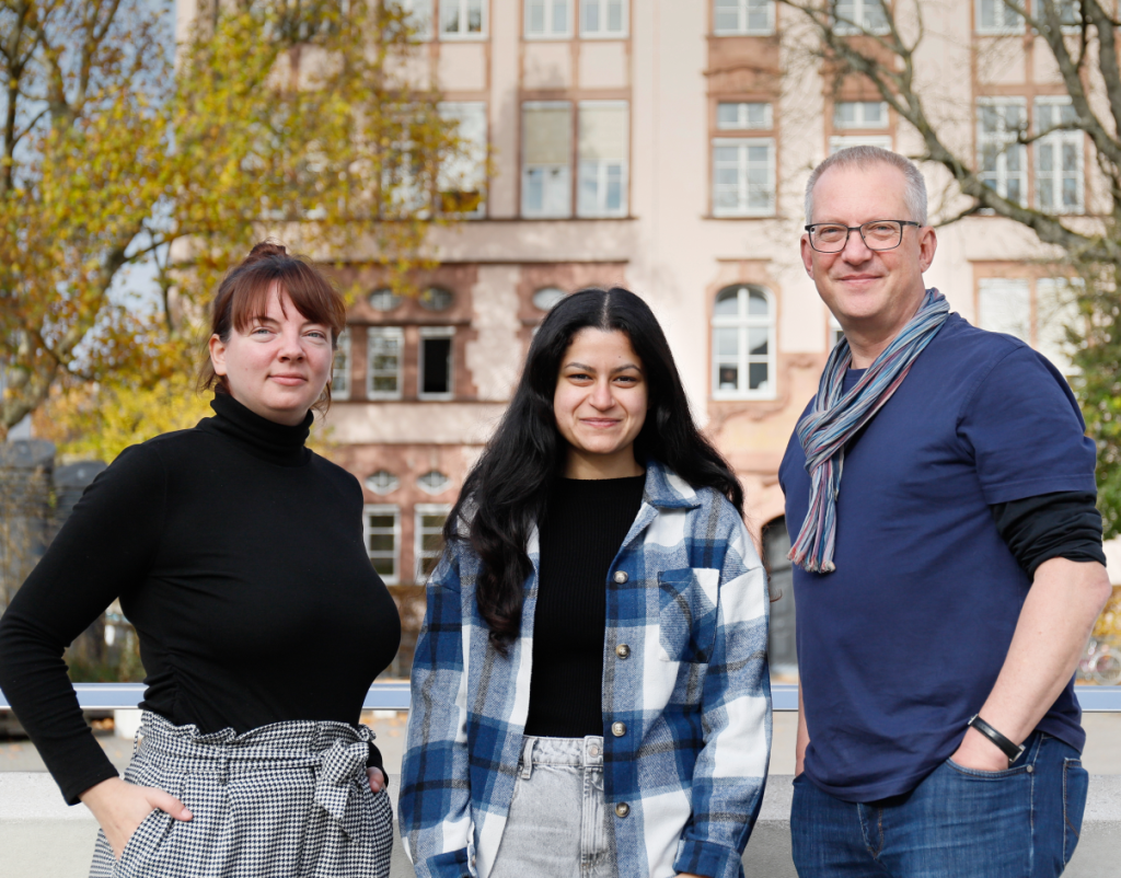Drei Erwachsene stehen nebeneinander. Von links nach rechts eine Frau mit schwarzem Oberteil und roten hochgesteckten Haaren, eine Frau mit Blau-weiß kariertem offenen Hemd und schwarzen Haaren und ein Mann mit blauem Shirt, Brille und kurzen brauen-grauen Haaren.