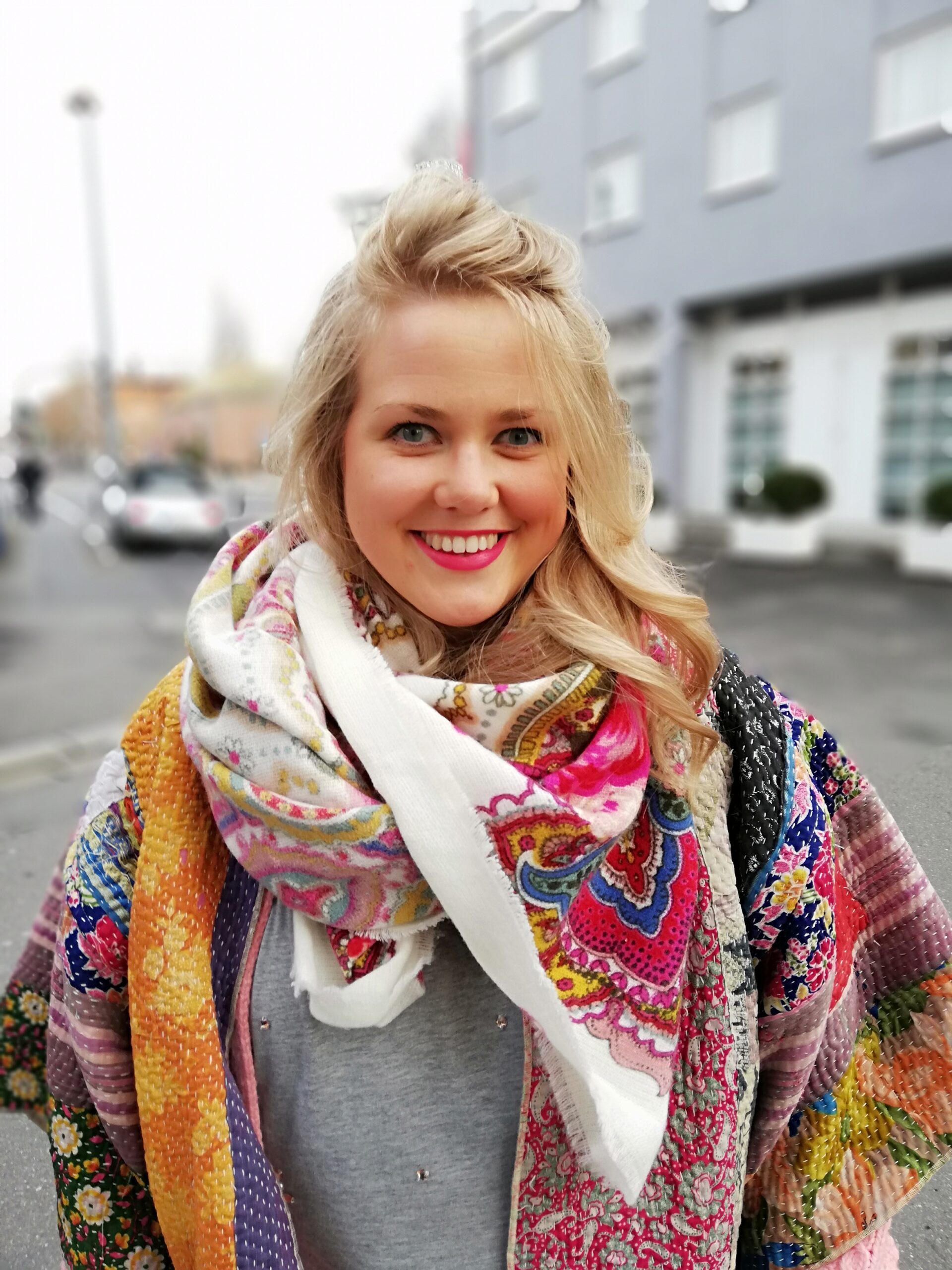 Ein Bild einer ca. 40 Jahre alten Frau mit blonden Haaren und. Sie lächelt in die Kamera. Ihre Jacke ist bunt und ihre Hallstuch breit mit der Grundfarbe weiß und weiteren bunten Farben. Sie steht in einer Einfahrt.