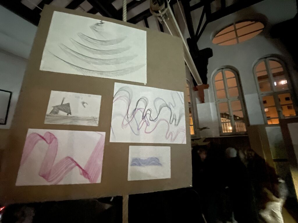 Bilder mit vielen Strichen hängen an einer Pinnwand. die in der Turnhalle steht. Das Licht ist dunkel und nur wenig zu erkennen.