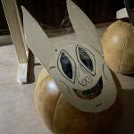 Eine Make aus Pappe mit großen ovalen länglichen Augen, langen spitzen Ohren, einem großen breiten Grinsen liegt auf einem großen Medizin-Sportball