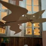 Ein brauner Pappdinosaurier fliegt an dünnen Fäden befestigt durch die Turnhalle. Im Hintergrund ist der nächtliche Himmel orange gefärbt.