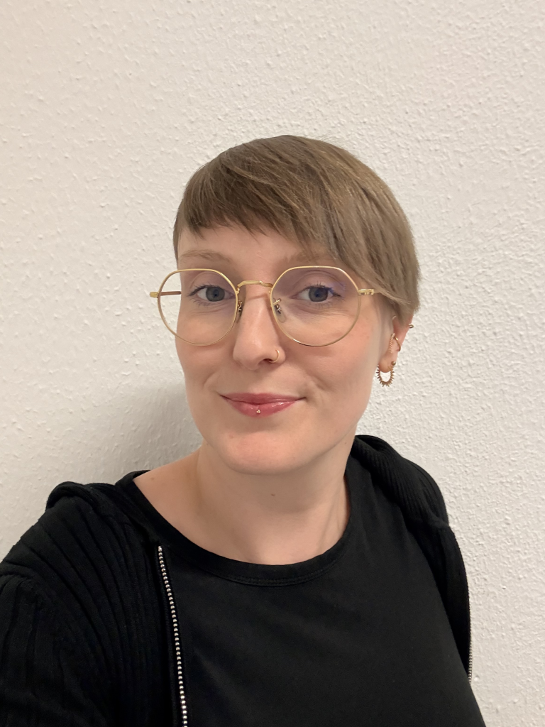 Eine Frau mit kurzen dunkelblonden Haaren, einer Brille mit hellem Rand und einem schwarzen Shirt.