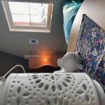 Eine weiße orientalisch anmutende Lampe im Vordergrund steht auf einem runden Tisch mit einer lila gemusterten Decke. Im Hintergrund ein Fenster und ein hellblauer Sitzsack