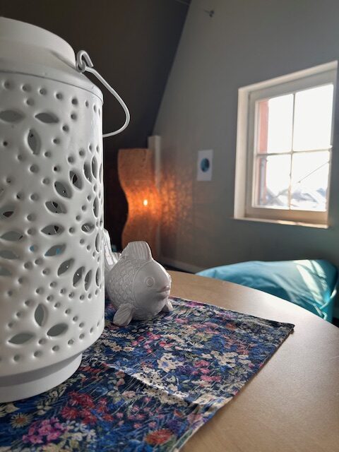Eine weiße orientalisch anmutende Lampe im Vordergrund steht auf einem runden Tisch mit einer lila gemusterten Decke. Im Hintergrund ein Fenster und ein hellblauer Sitzsack