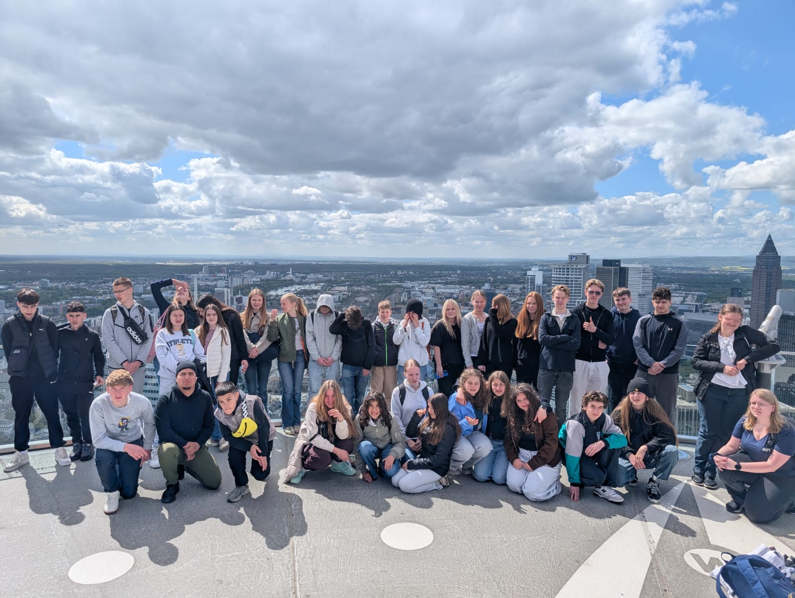Eine große Gruppe von Jugendlichen und Erwachsenen stehen und hocken zusammen auf der Aussichtsplattform des Main-Towers. Die Wolken sind klar aber auch grau.