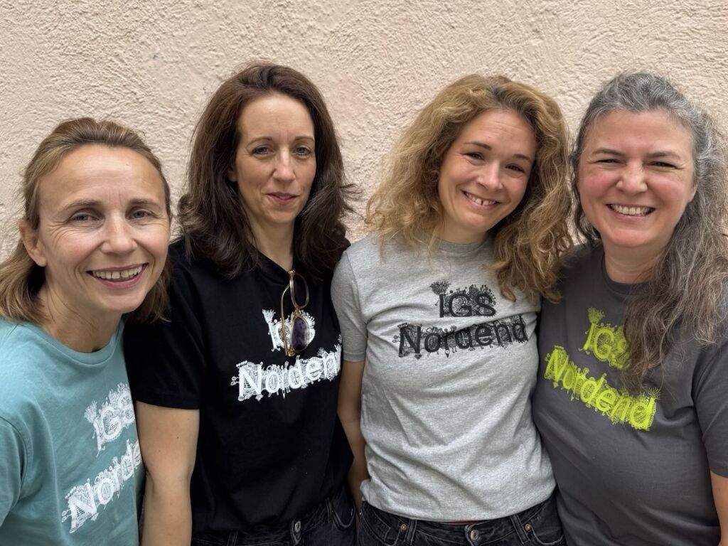 Vier Frauen stehen eng nebeneinander . Rechts mit marinblauem, daneben mit schwarzem, den mit hellgrauen und ganz rechts mit dunkelgrauem T-Shirt mit einem verschnörkelten Schriftzug "IGS Nordend".