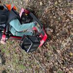 Laub auf dem Rasen des Gartens, ein Rechteck mit Faben abgesteckt, ein roter Rollstuhlbuggy mit Person mit lila Hose steht darauf.
