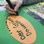 Eine Hand mit Stift beschreibt eine ovale orange Moderationskarte mit Der GRüne Daumen